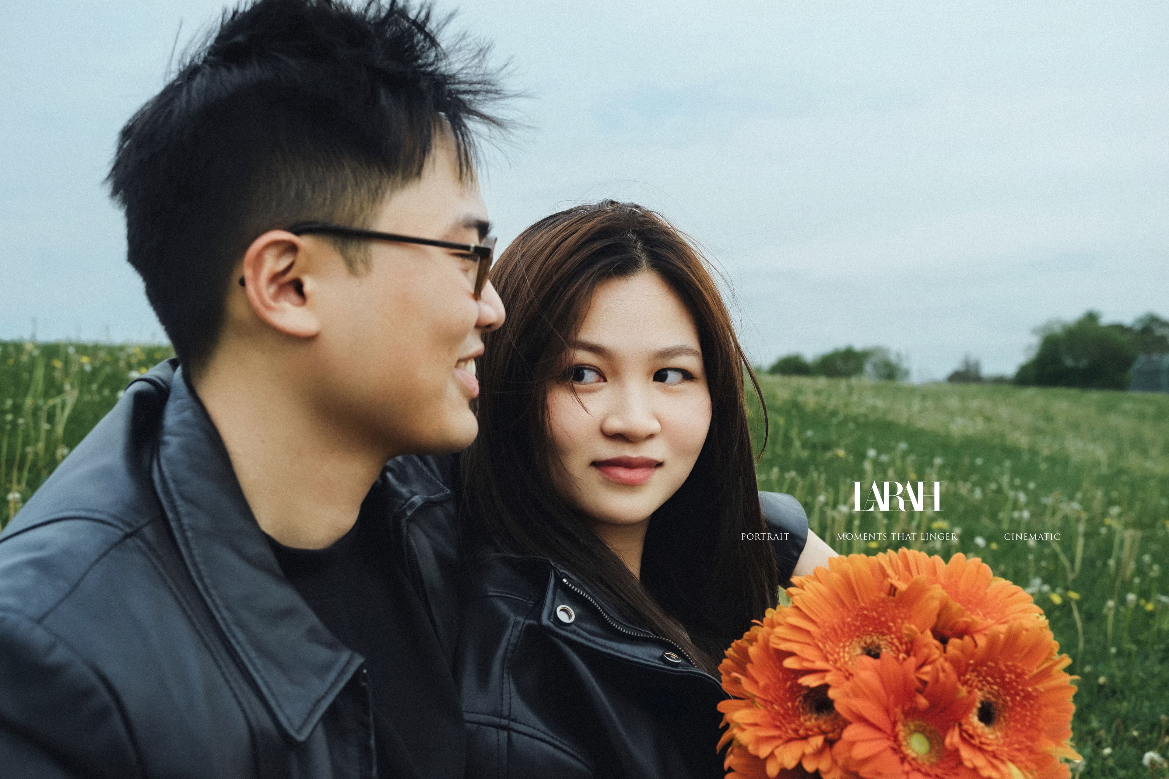 Couple standing back to back in an open meadow, both in black leather jackets, the woman holding orange gerbera daisies with her eyes closed.
