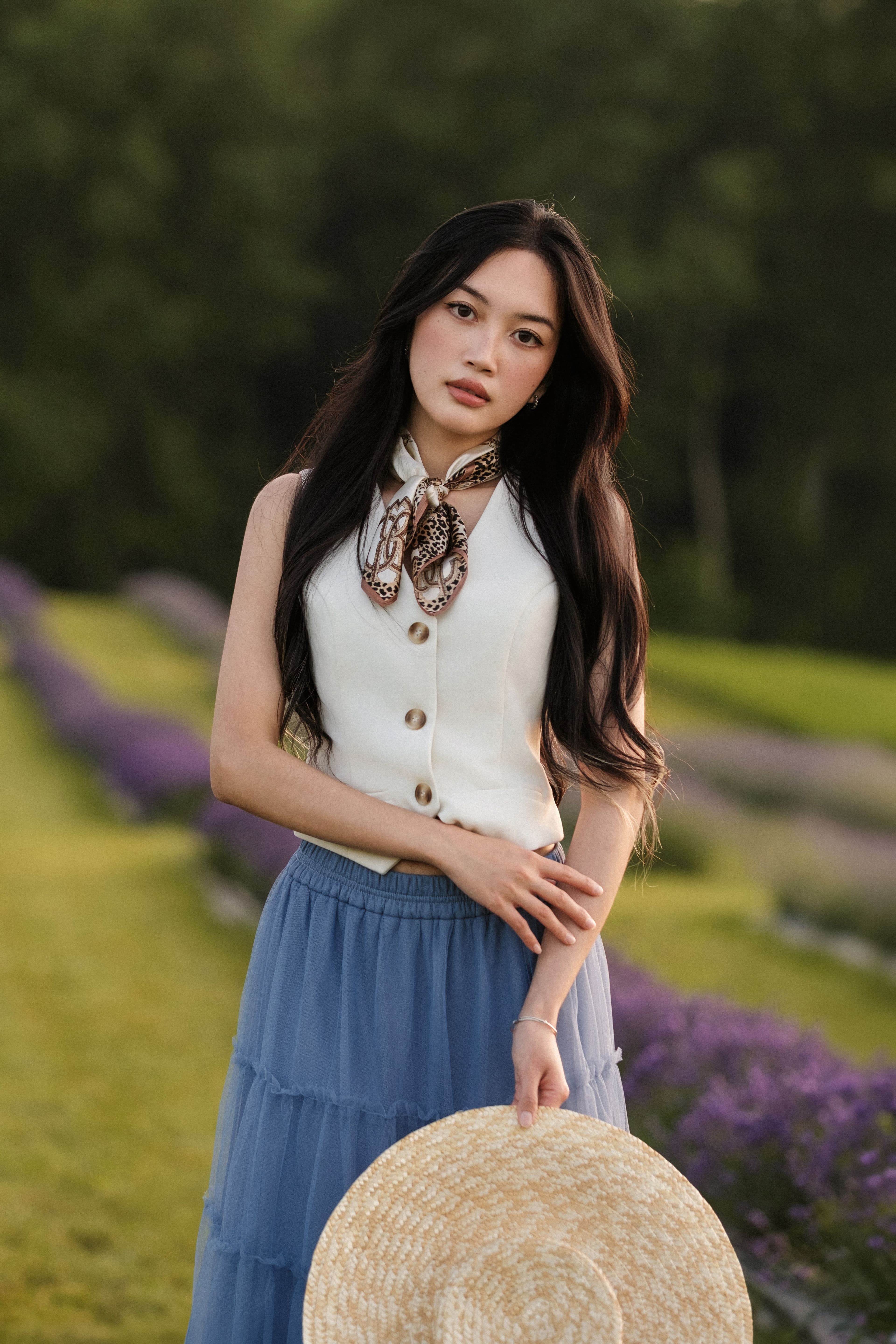 Woman standing in a lavender field holding a wide straw hat at her side.