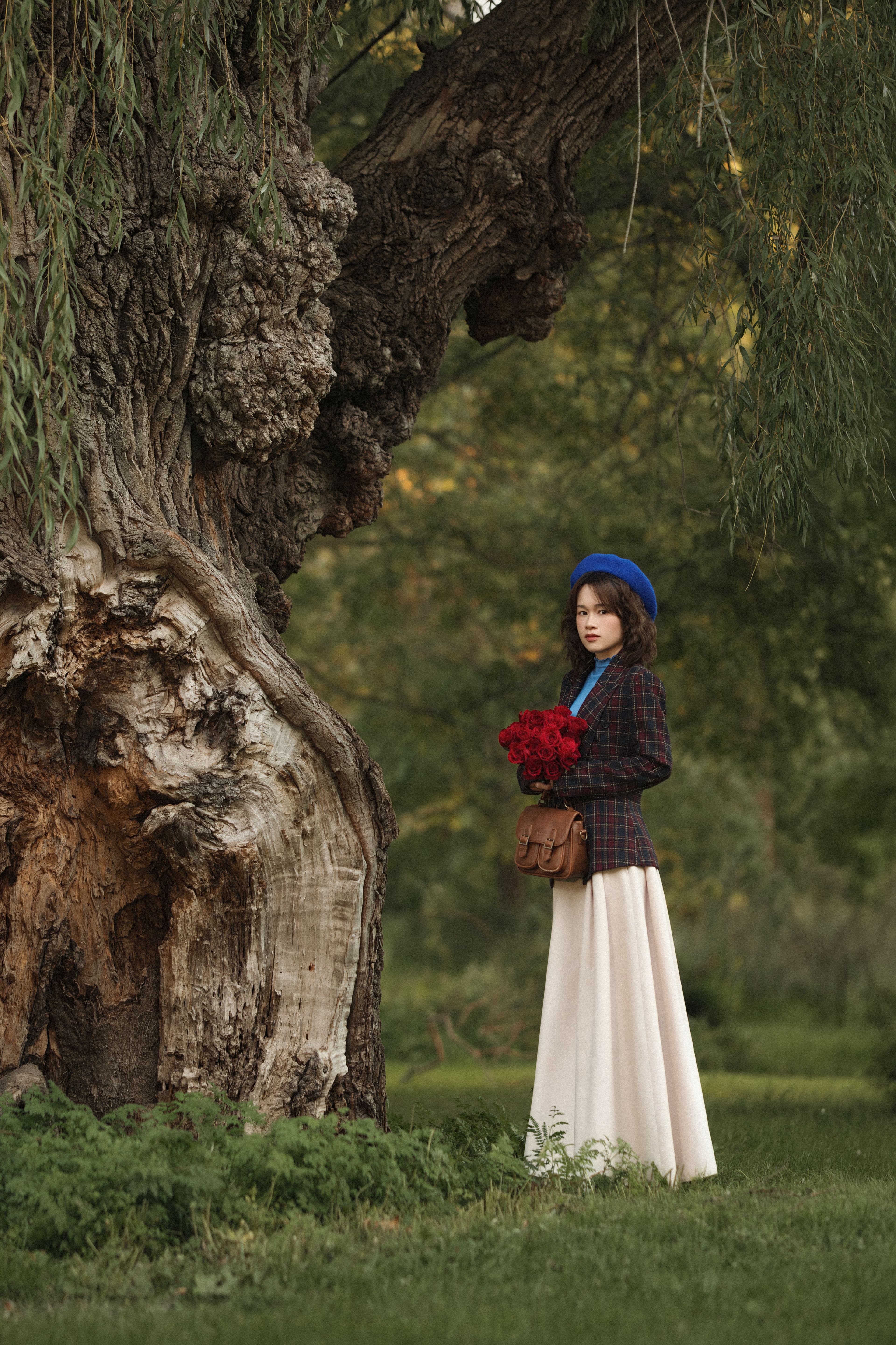Woman in a blue beret and plaid blazer holding red roses beside an old willow tree.